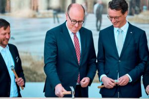 Ministerpräsident Wüst und Staatssekretär Hauer schlagen den symbolischen letzten Nagel in den Balken ein.