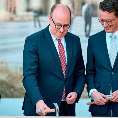 Ministerpräsident Wüst und Staatssekretär Hauer schlagen den symbolischen letzten Nagel in den Balken ein.